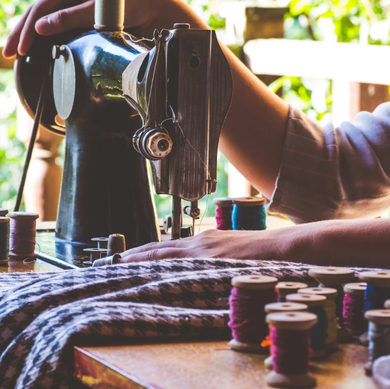 Person using a sewing machine with spools of thread on a table outdoors.